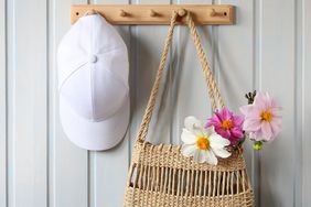 A wicker bag with a bouquet of dahlias and a white baseball cap hang on a wooden hanger on a board wall indoors.