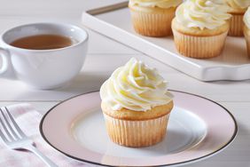 pale pink plate with cupcake with white frosting, cup of tea and tray of cupcakes